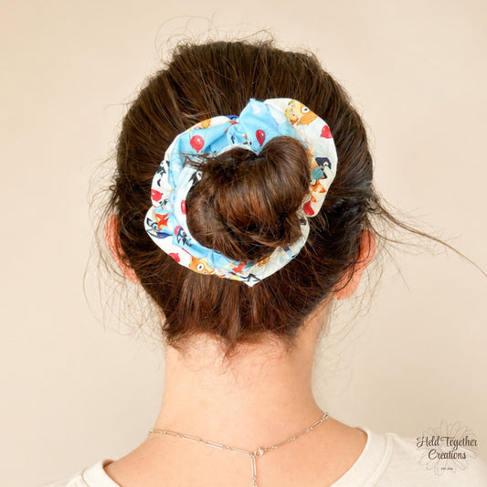 Balloon Bash Scrunchie worn around a brown messy bun, featuring a blue and white cartoon pup and balloon print.