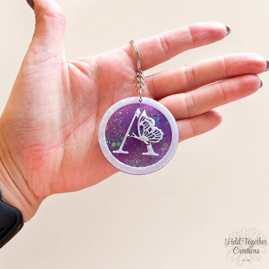 Hand holding a keychain with a purple and white design on a beige background