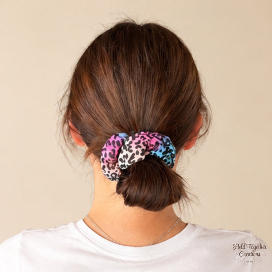 A person shown from the back wearing a leopard-print scrunchie in pink, blue, black, and white, tied around brown hair. The background is a solid light beige wall.
