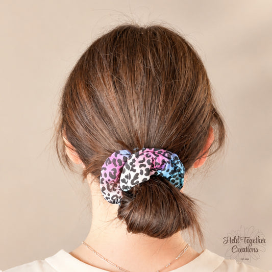 Colourful leopard print handmade scrunchie with pink, blue, purple and black tones on a soft ivory background.