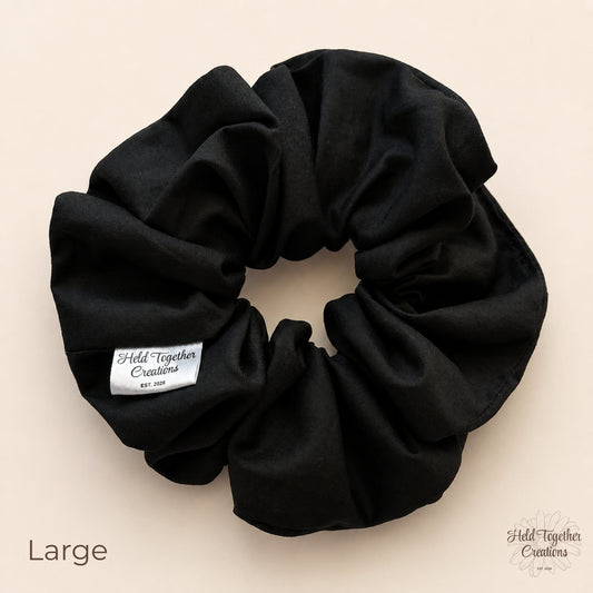 A large black fabric scrunchie with a white brand label reading 'Held Together Creations' on a light beige background. The word 'Large' is printed in the lower-left corner, and the brand logo appears in the lower-right corner.