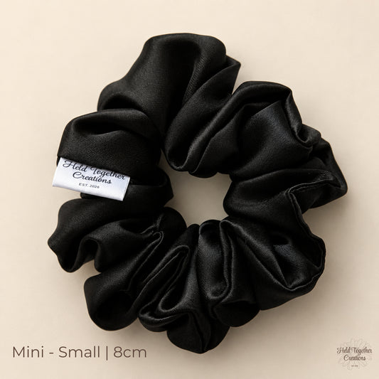 Black satin Mini scrunchie (8cm) laid flat on a warm ivory background, showing soft gathered folds and Held Together Creations label.