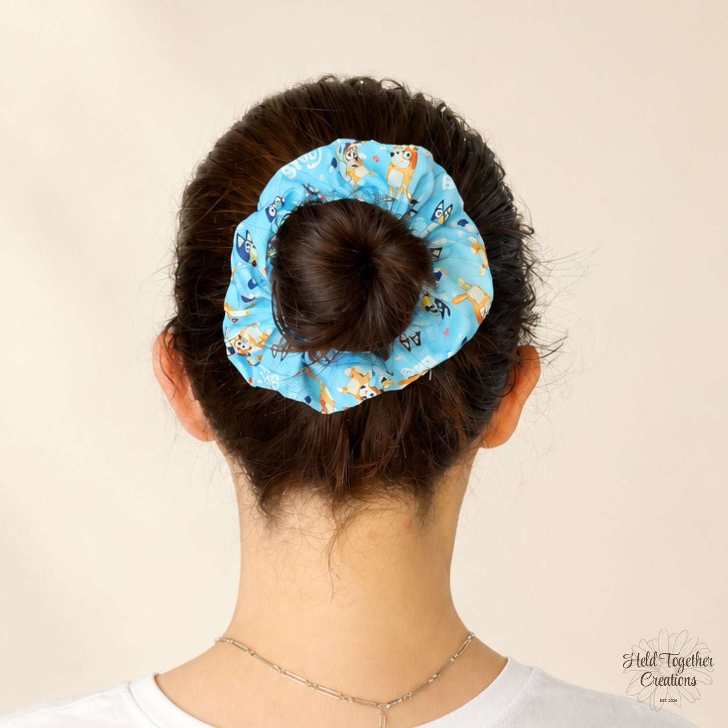 A light blue fabric scrunchie with a cartoon dog pattern, shown on a model's hair in a bun from the back against a plain beige background.