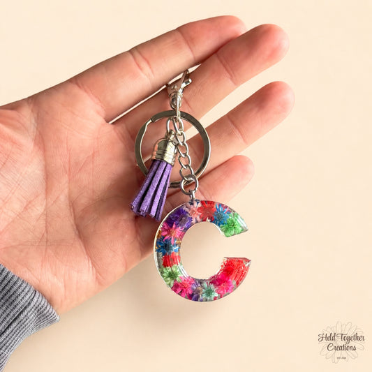 A hand holding a keychain with a colourful floral letter C charm, a purple tassel, and silver metal rings on a light beige background.