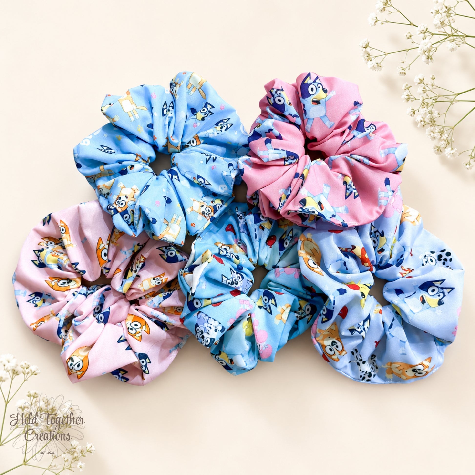 Backyard Besties handmade scrunchies in colourful blue and pink puppy-print fabrics on a warm ivory background with delicate baby’s breath flowers.