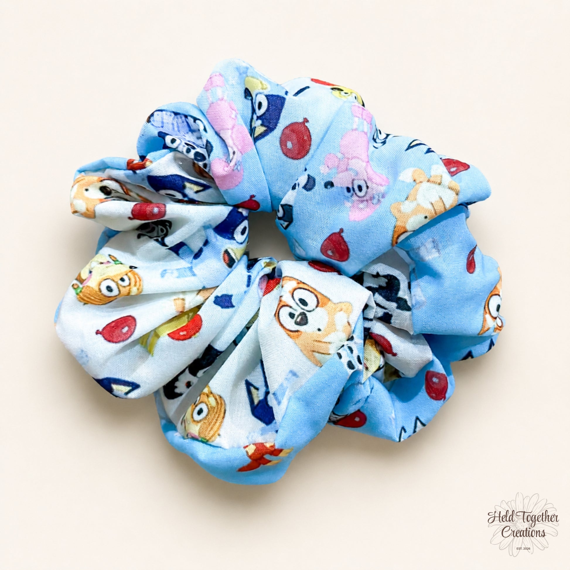 A fabric hair scrunchie featuring a light blue background with a colourful cartoon animal print, including a bingo, a coco, and red balloons. The scrunchie is photographed on a plain, warm off-white background.