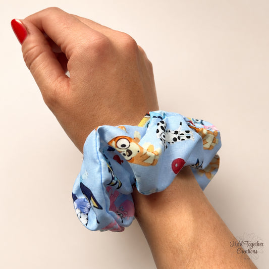 A light blue fabric hair scrunchie with a cartoon animal print, shown worn on a person's wrist. The print features orange, black, and white cartoon characters.