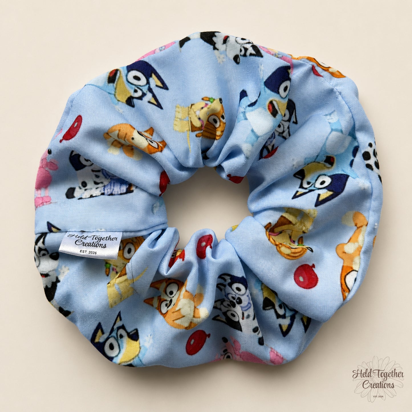 A light blue fabric scrunchie with a colorful cartoon animal print, featuring a white fabric brand tag that reads 'Field Together Creations Est. 2026' and a matching watermark in the bottom right corner.