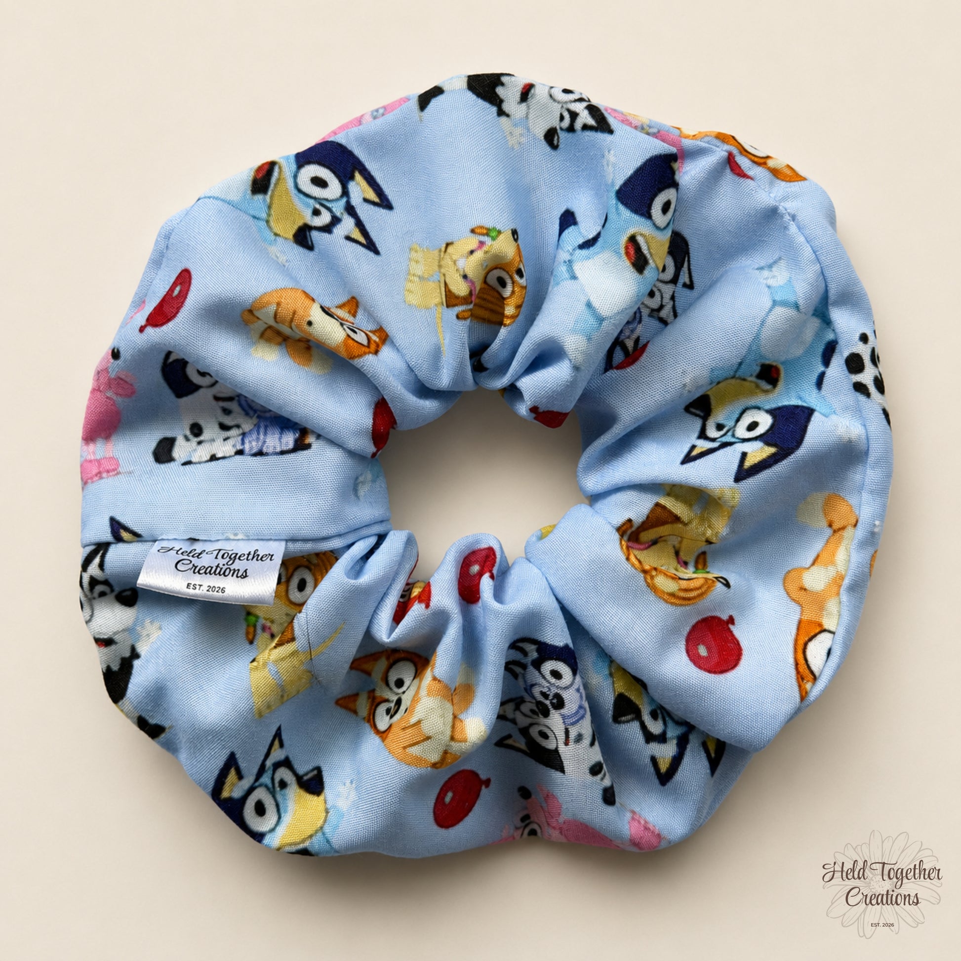 A light blue fabric scrunchie with a colorful cartoon animal print, featuring a white fabric brand tag that reads 'Field Together Creations Est. 2026' and a matching watermark in the bottom right corner.