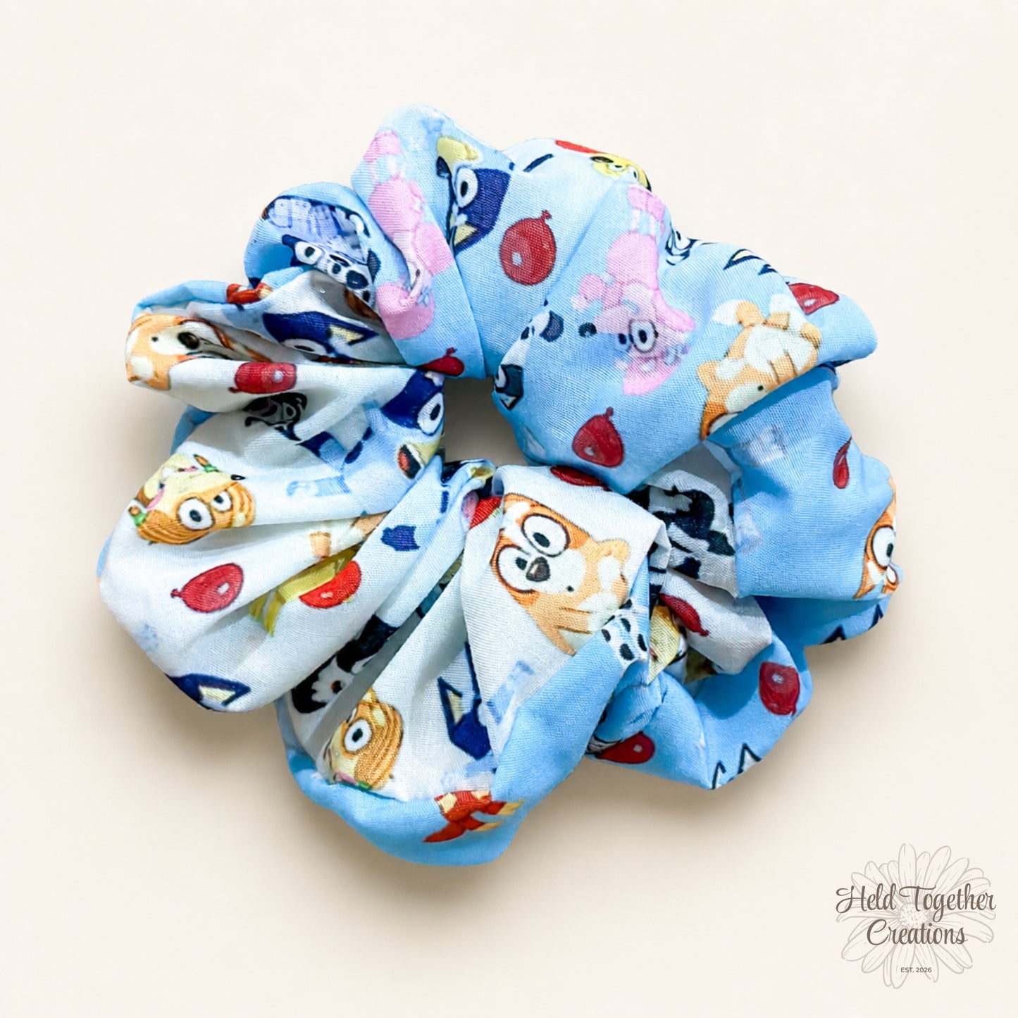 Balloon Bash handmade scrunchie in light blue puppy-print fabric on a warm ivory background with Held Together Creations branding.