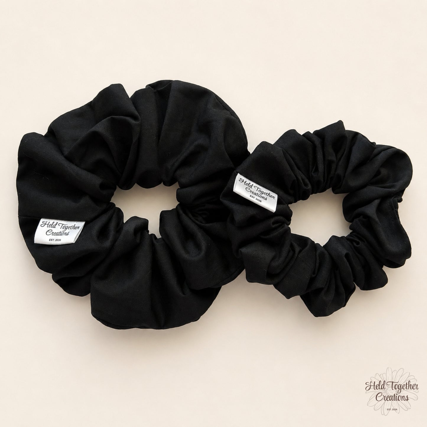 Small and large black cotton scrunchies displayed together on a warm ivory background.