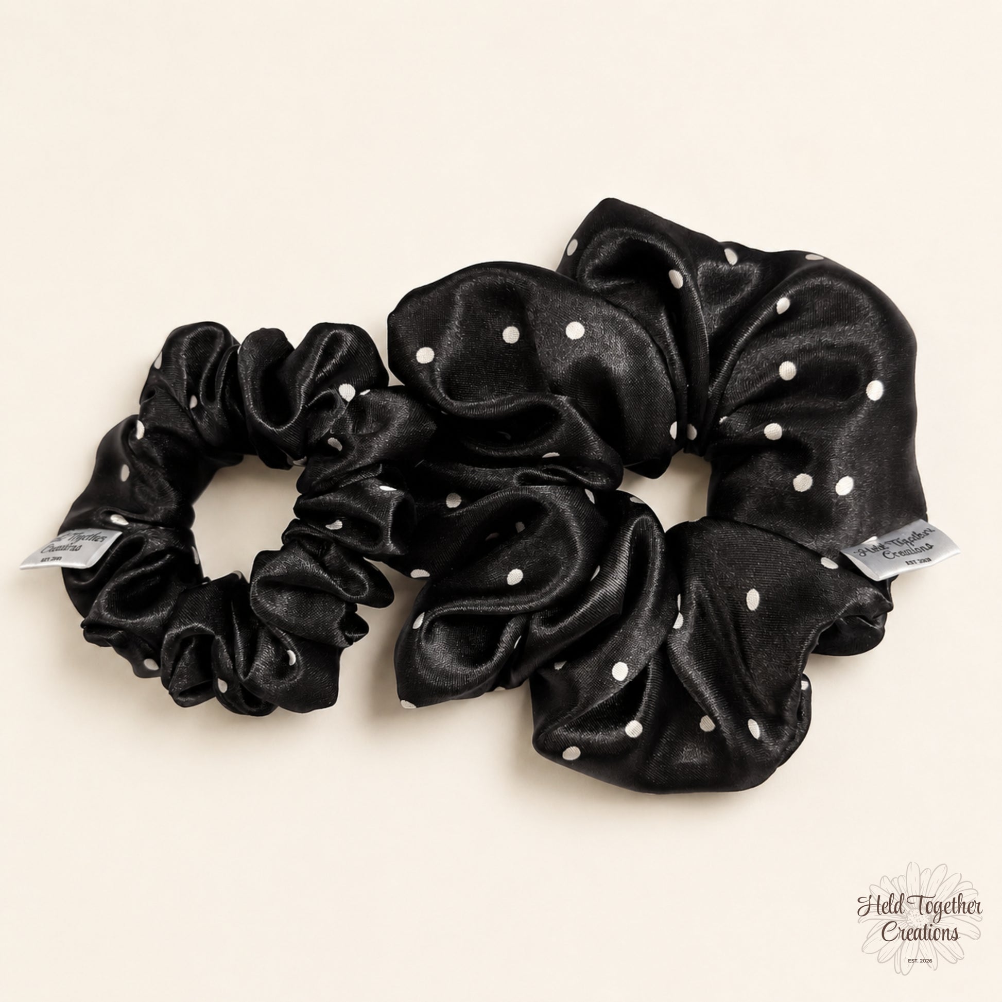 Small and large black satin scrunchies with white spot pattern displayed together on a soft ivory background.