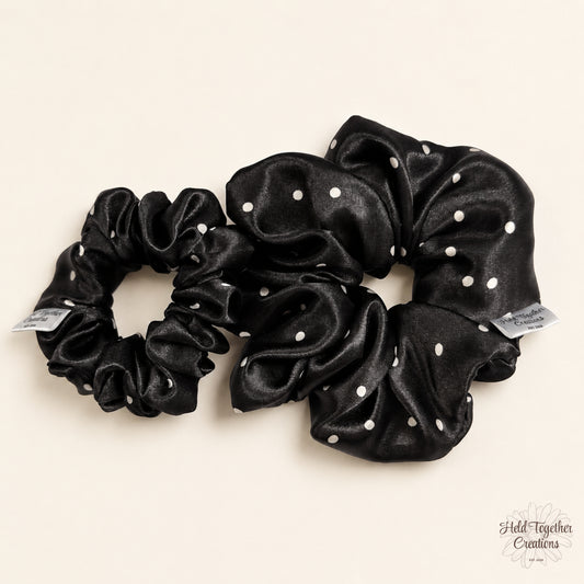 Small and large black satin scrunchies with white spot pattern displayed together on a soft ivory background.