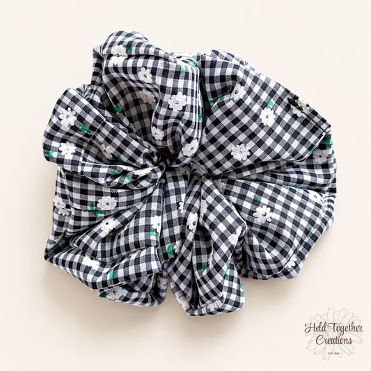 Black and white gingham floral fabric scrunchie displayed flat on a warm ivory background.