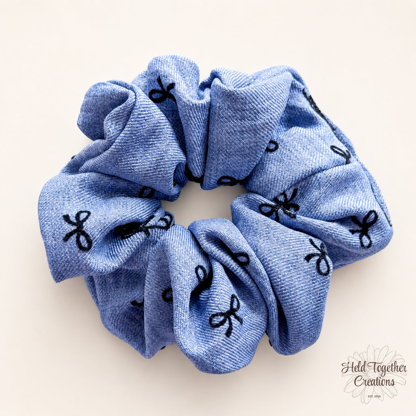 Blue handmade fabric scrunchie with black bow pattern on a warm ivory background.