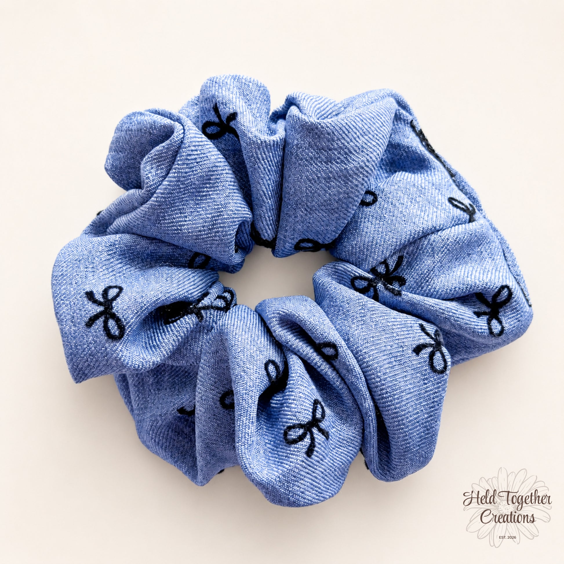 Blue handmade fabric scrunchie with black bow pattern on a warm ivory background.