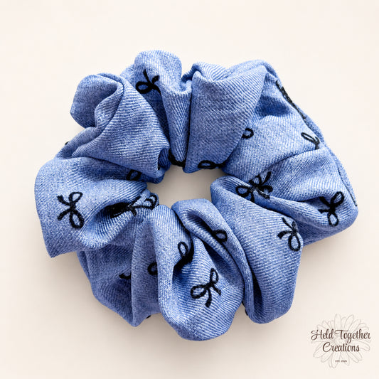Blue handmade fabric scrunchie with black bow pattern on a warm ivory background.