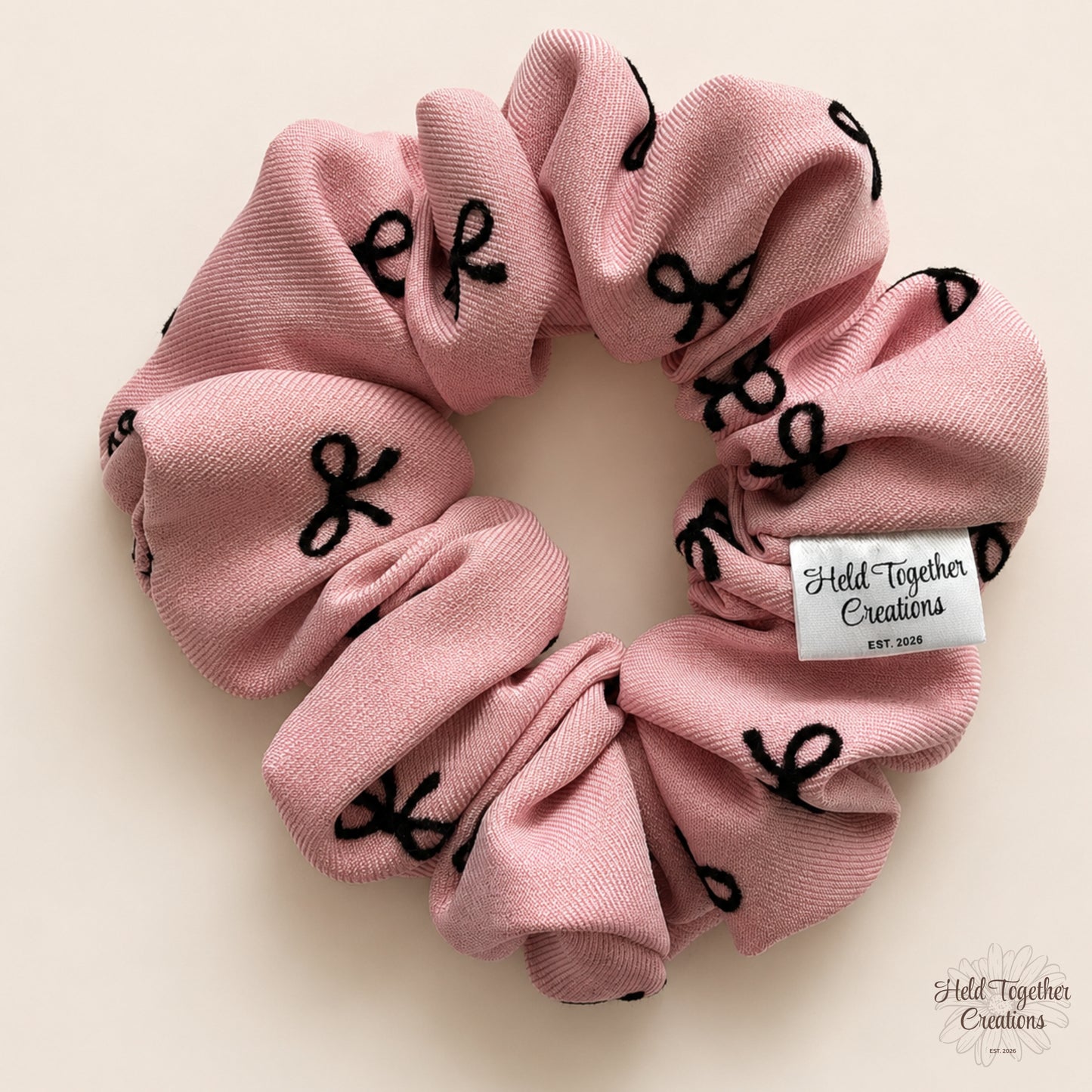 Blush pink fabric scrunchie with scattered black bows displayed flat on a warm ivory background.