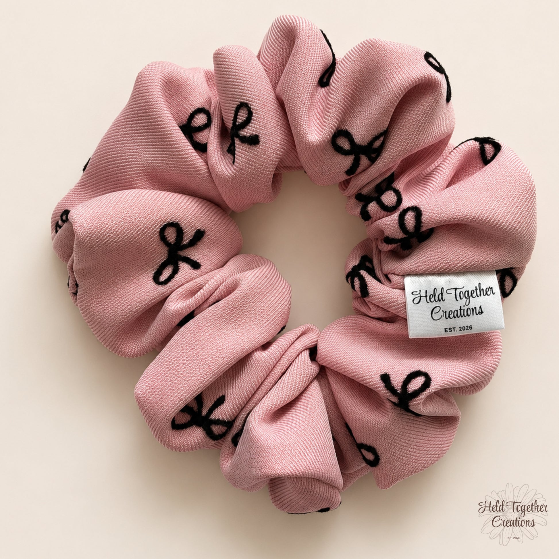 Blush pink fabric scrunchie with scattered black bows displayed flat on a warm ivory background.