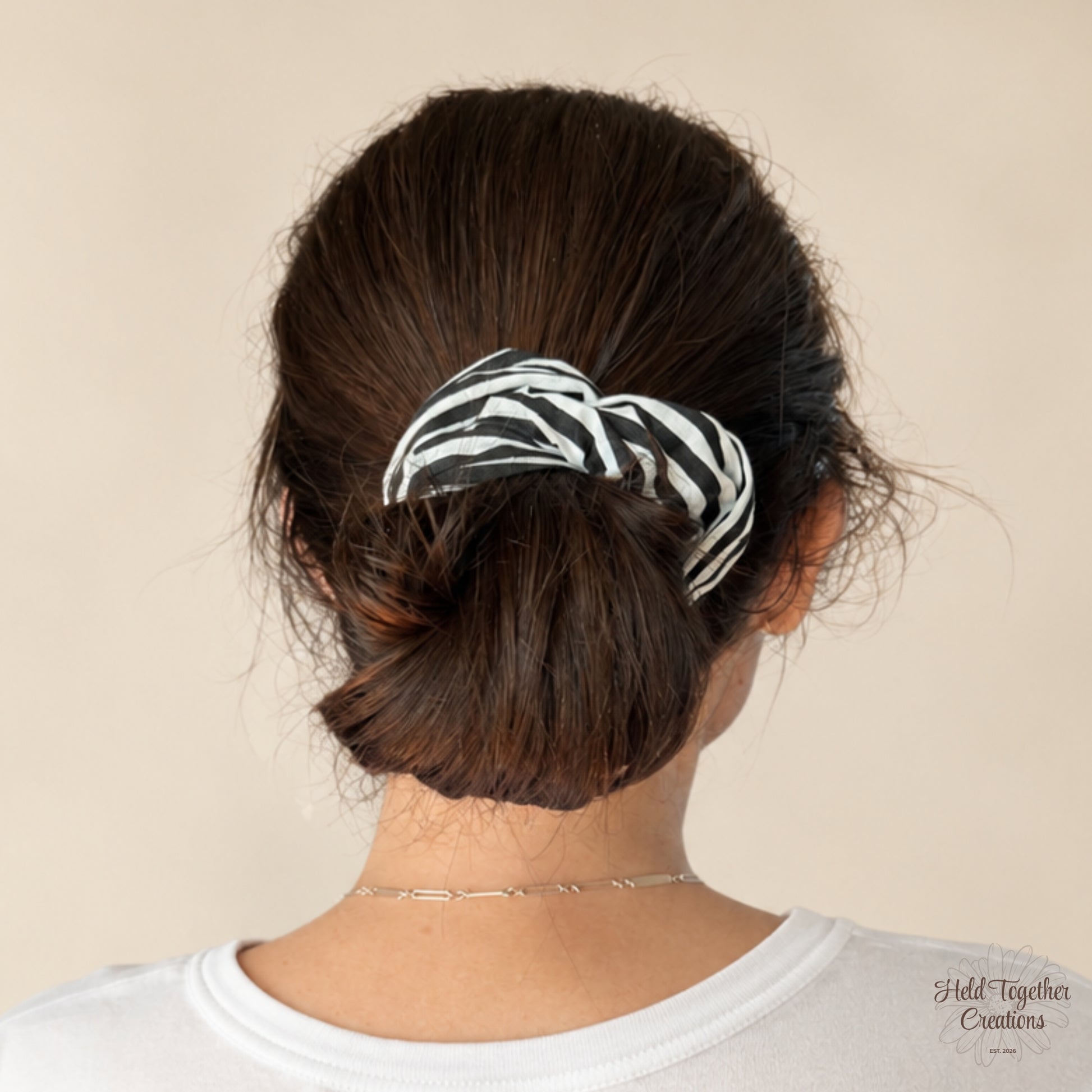 A woman viewed from the back wearing a black and white scrunchie to hold her dark brown hair in a low bun, against a plain beige background.