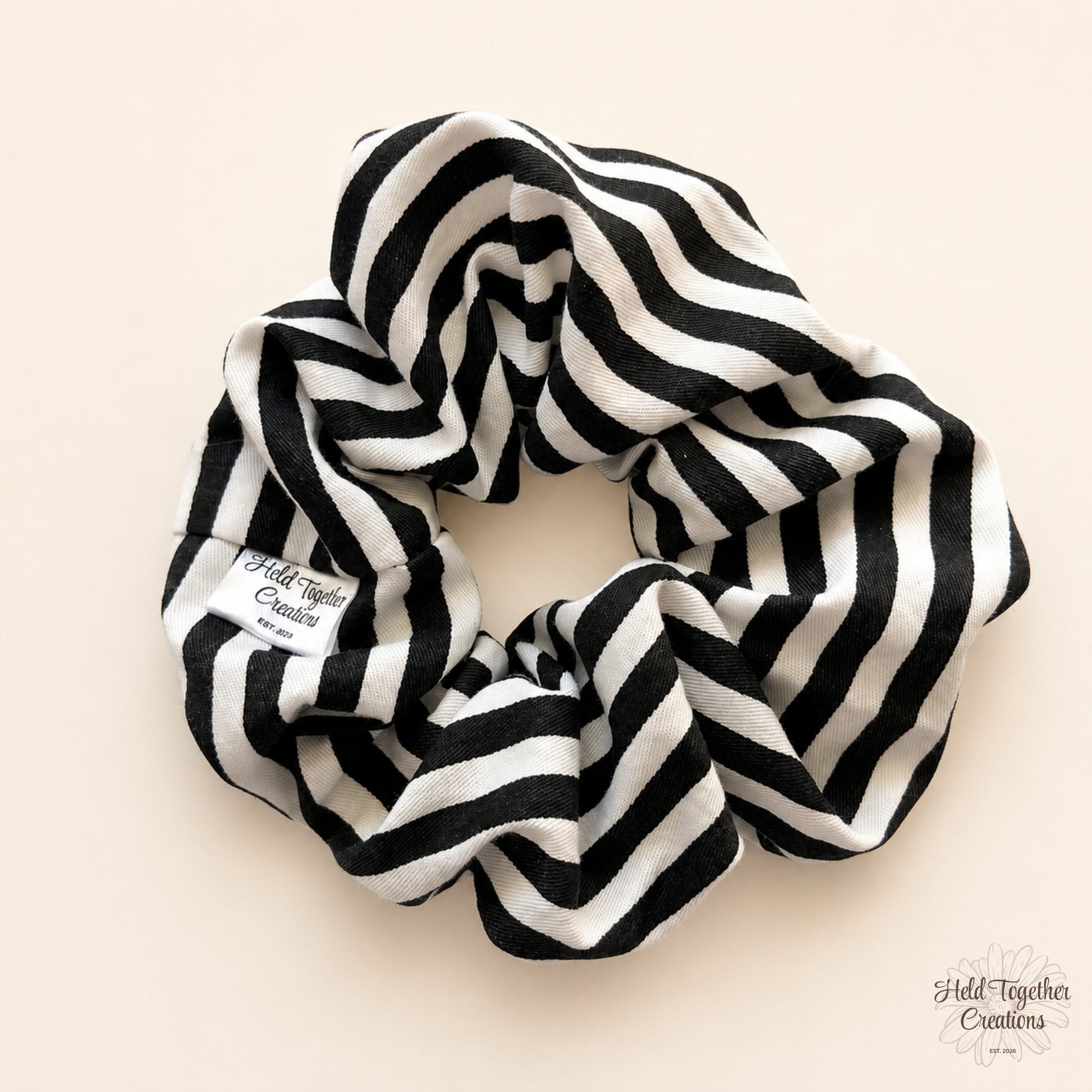 A fabric hair scrunchie with a black and white wavy stripe pattern, displayed on a light beige background. A small white fabric tag on the left side reads 'Held Together Creations'.