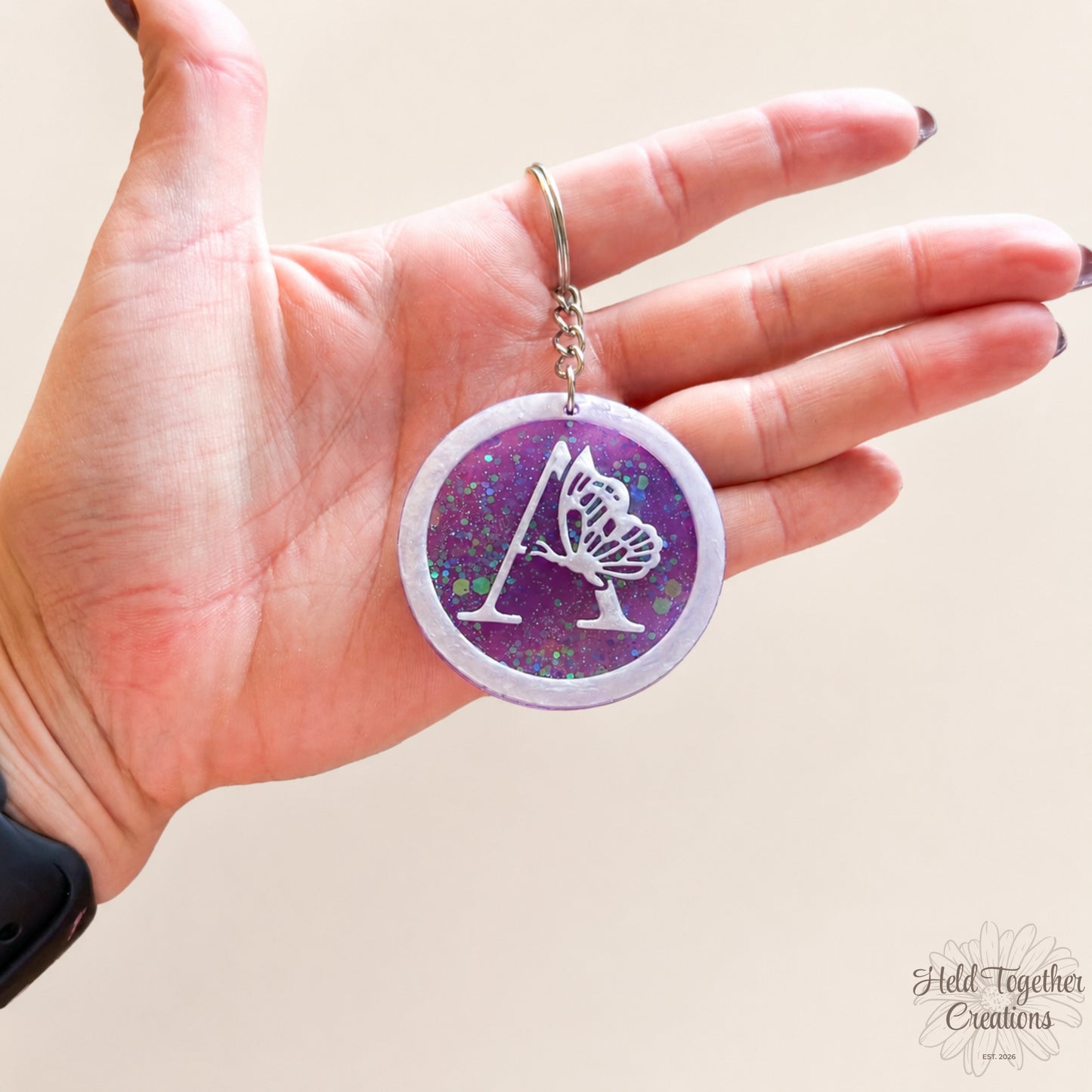 Hand holding a keychain with a purple and white design on a beige background