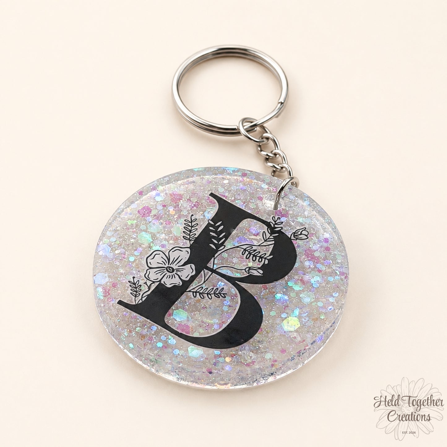 Handmade clear glitter resin initial bag tag with black floral letter B and silver keyring hardware on a warm ivory background.