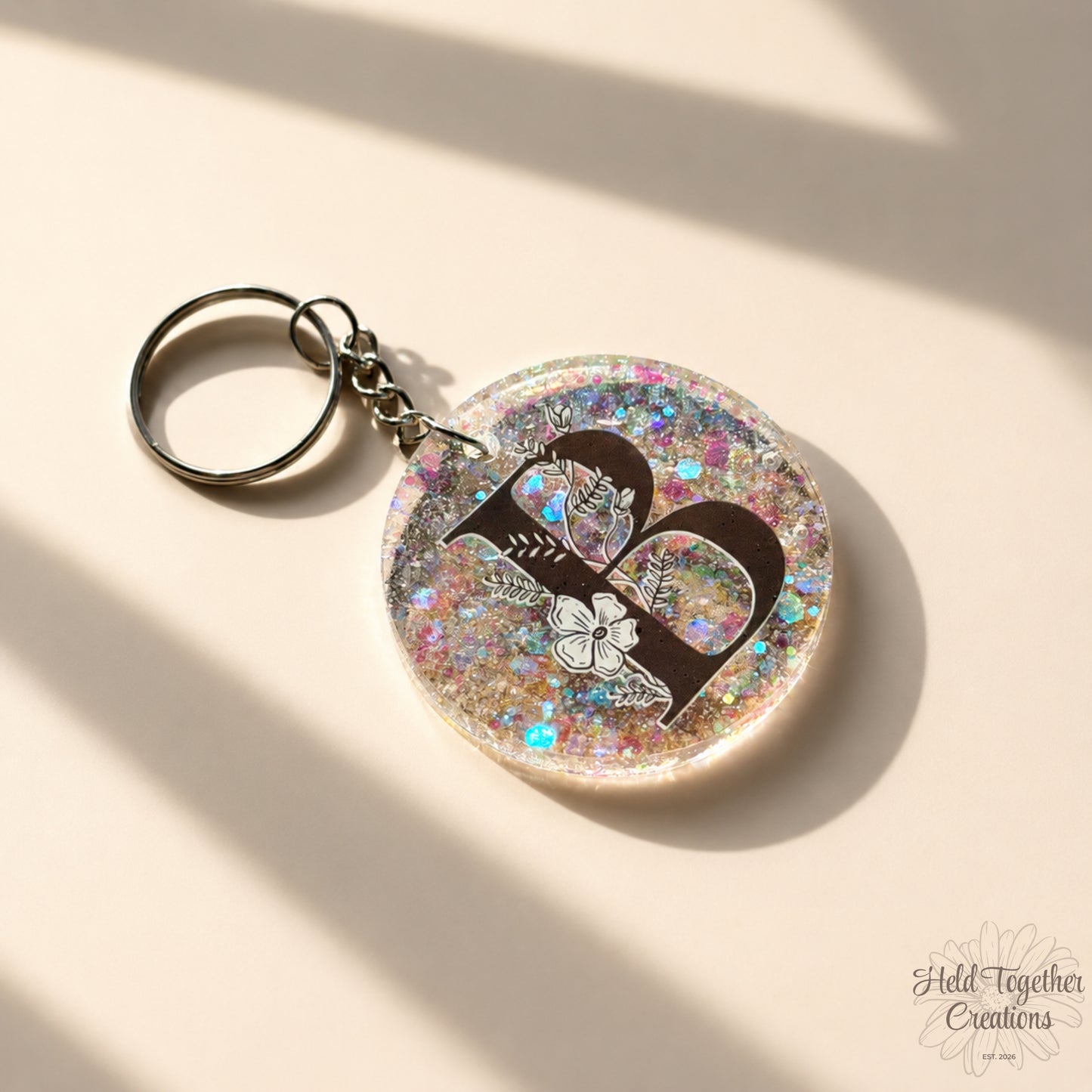 Handmade clear glitter resin initial bag tag with black floral letter B and silver keyring hardware on a warm ivory background.