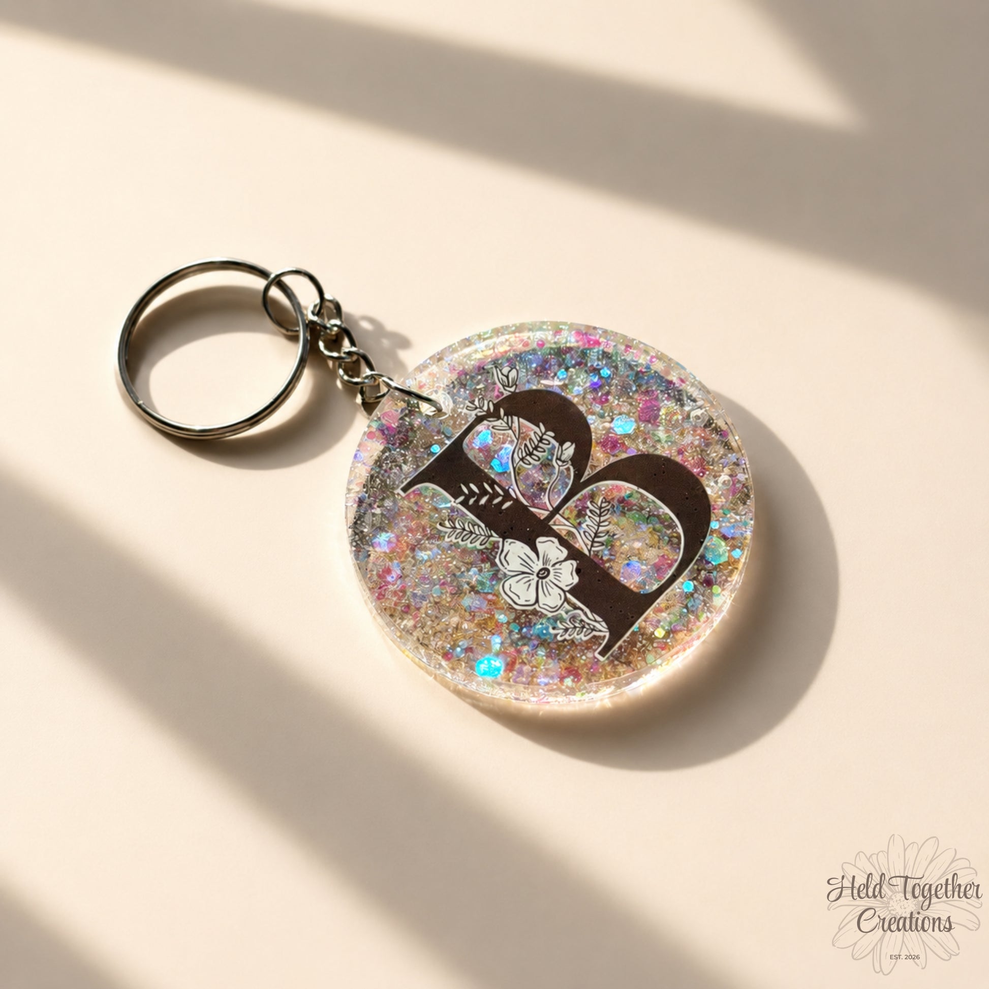 Handmade clear glitter resin initial bag tag with black floral letter B and silver keyring hardware on a warm ivory background.