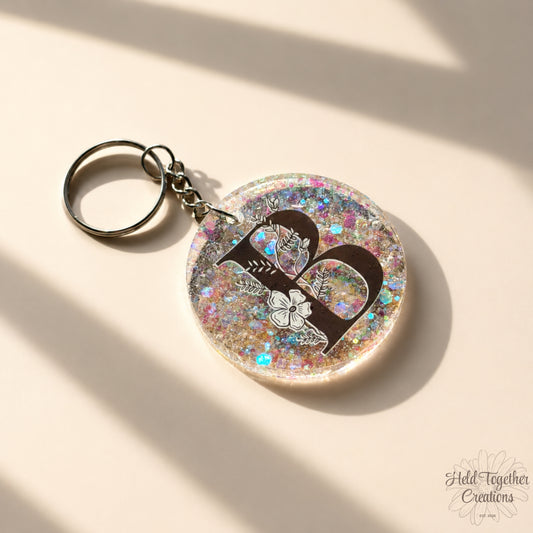 Handmade clear glitter resin initial bag tag with black floral letter B and silver keyring hardware on a warm ivory background.
