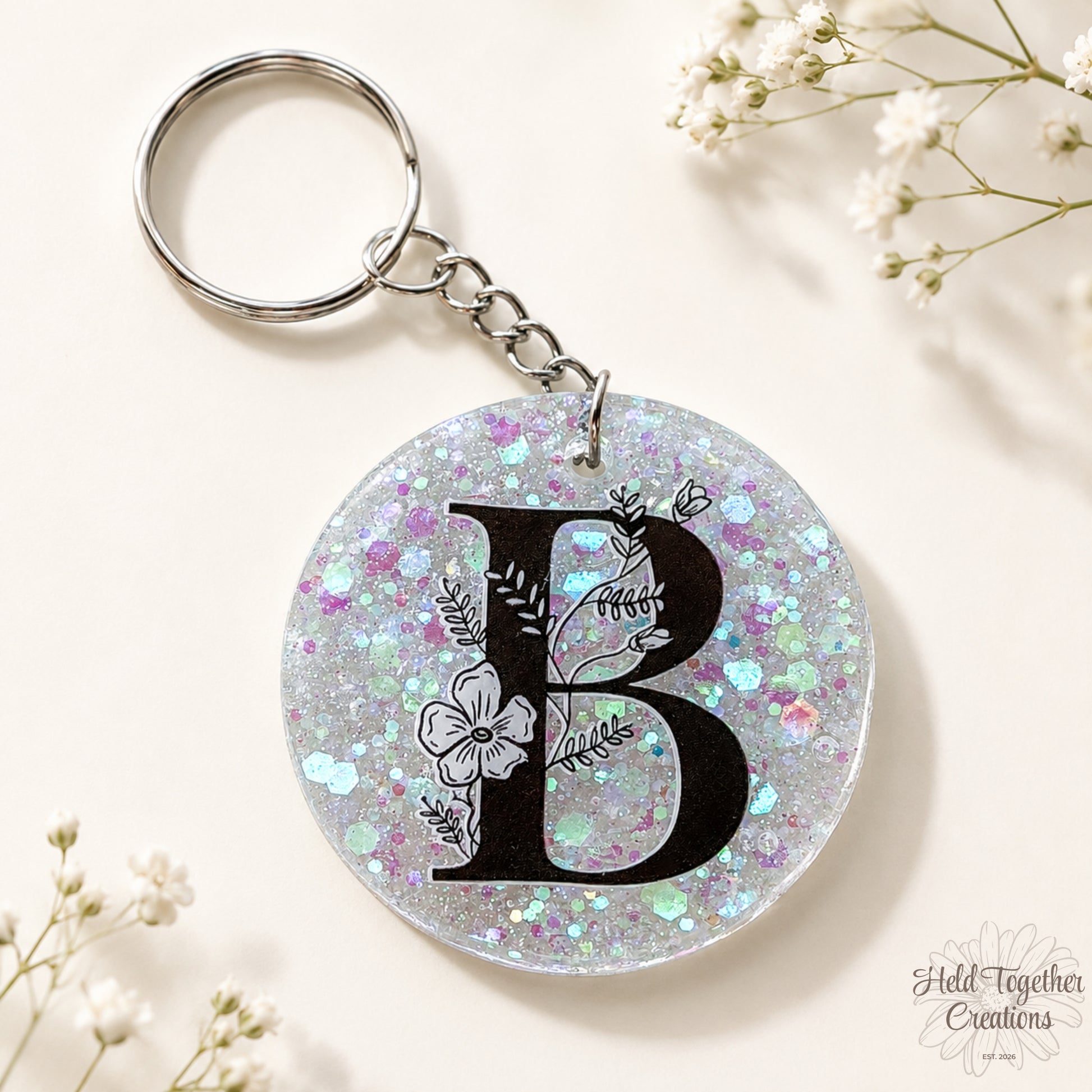 Handmade clear glitter resin initial bag tag with black floral letter B and silver keyring hardware on a warm ivory background with baby’s breath.