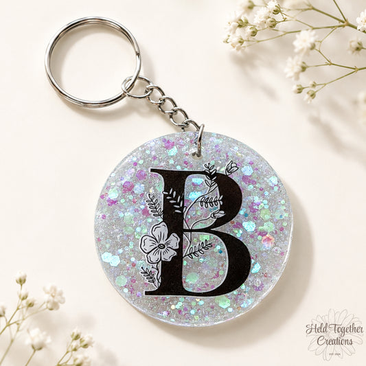 Handmade clear glitter resin initial bag tag with black floral letter B and silver keyring hardware on a warm ivory background with baby’s breath.