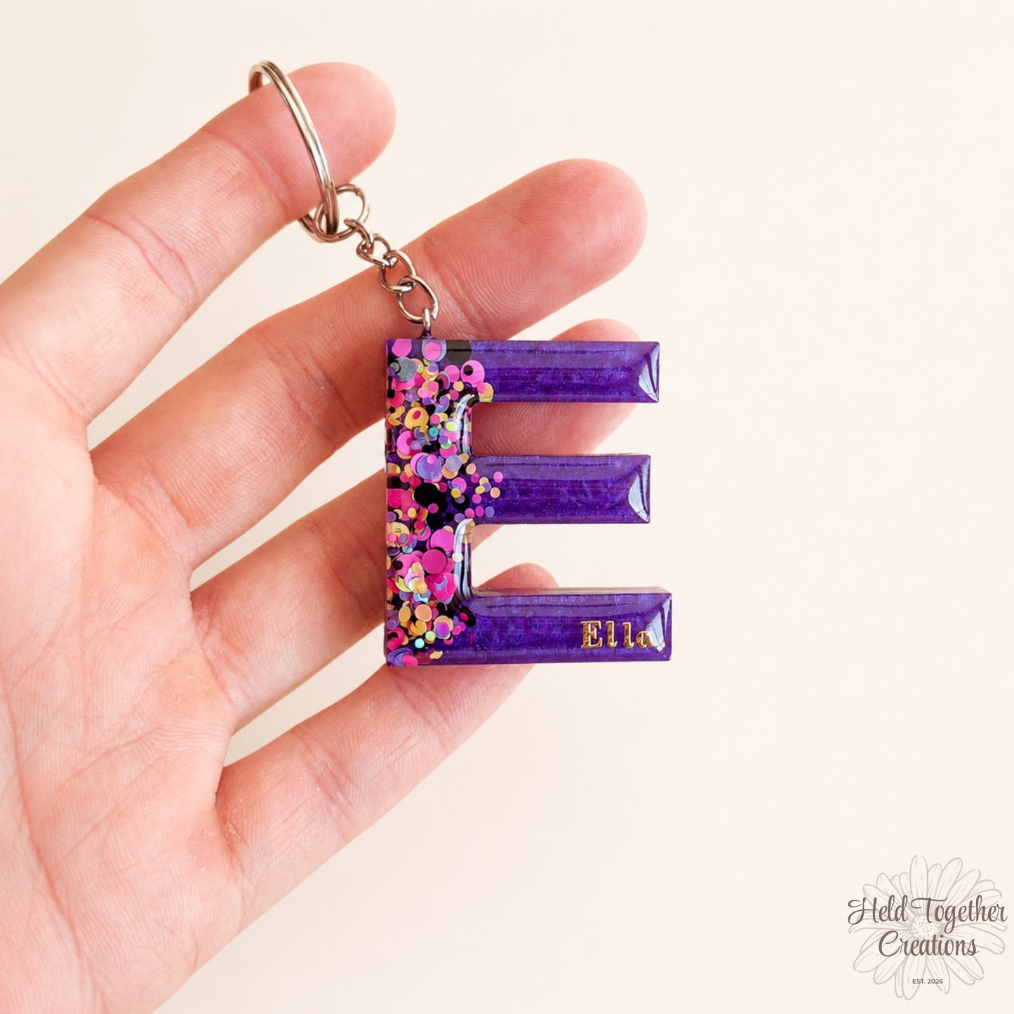 Hand holding a purple keychain with floral design and 'Ella' text on a light background