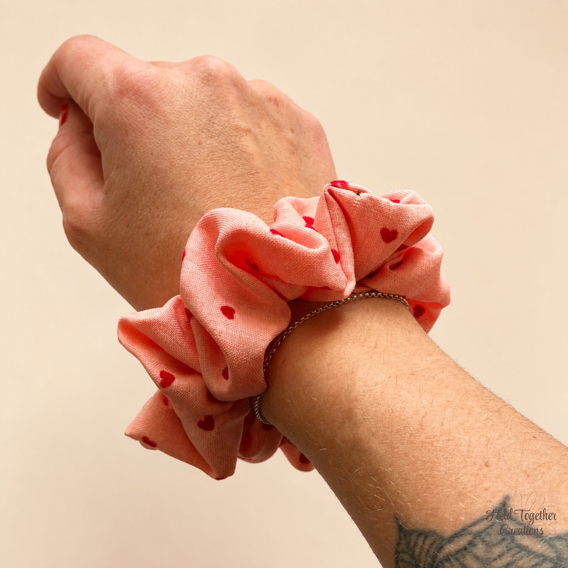 A close-up of a person's wrist wearing a coral pink fabric scrunchie with small red hearts, shown against a plain beige background.