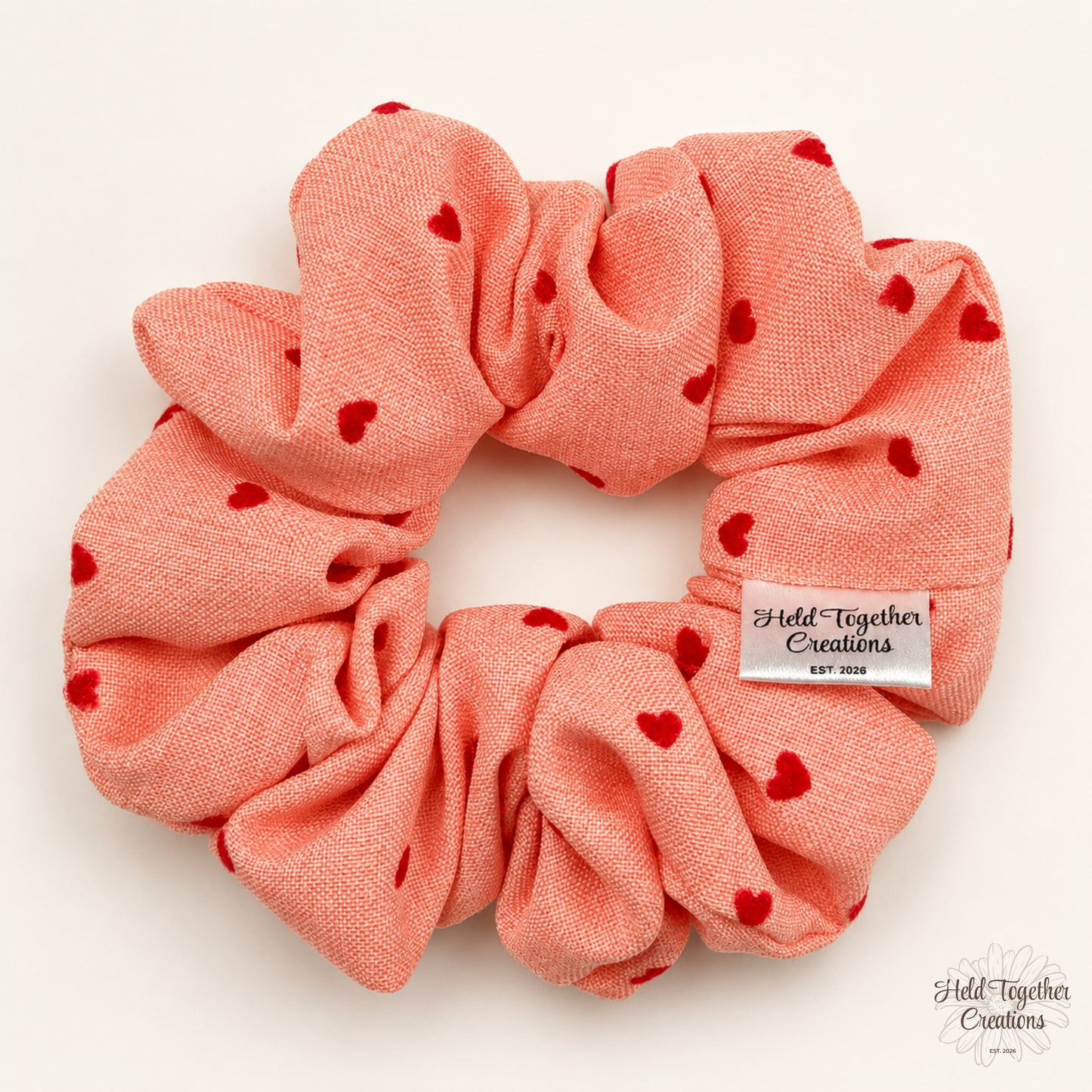 A coral pink fabric scrunchie with small red heart prints, featuring a white brand tag that reads 'Held Together Creations'.