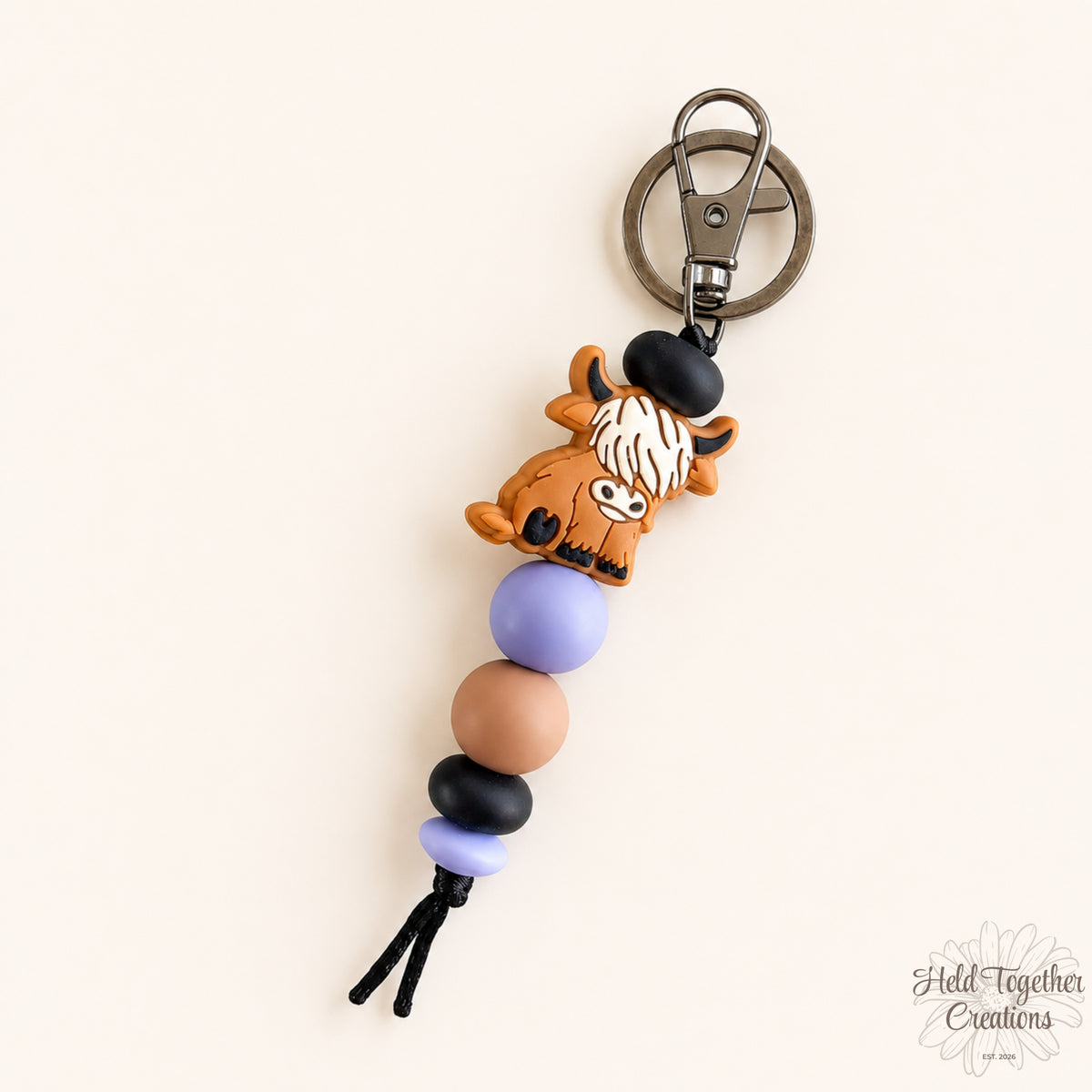 Purple Cosy Cow silicone bead keyring with a brown highland cow charm, lilac, tan and black beads on a warm ivory background.
