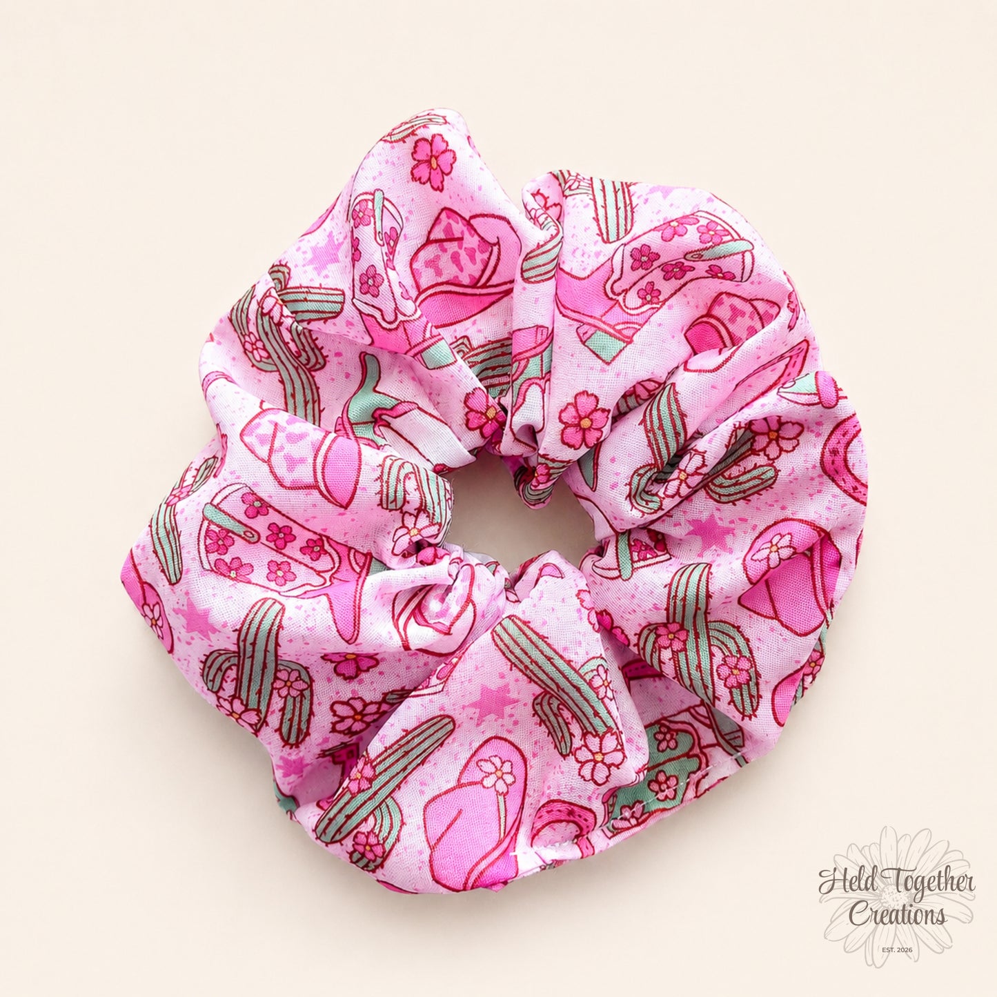 Pink Cowgirl Crush handmade scrunchie on a warm ivory background, featuring a playful western print with cowgirl boots, hats, cacti and flowers.