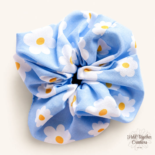Light blue handmade scrunchie with white daisy print and yellow flower centres on a soft neutral background.