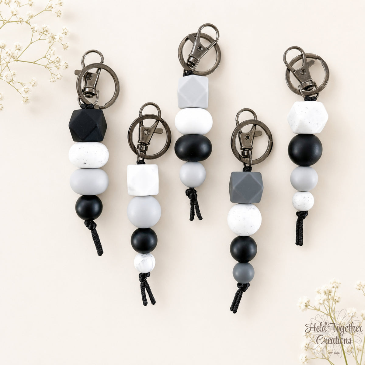 Group photo of five Dark & Stormy silicone bead keyrings in black, grey, white, speckled and marble-look designs on a warm ivory background with soft floral styling.