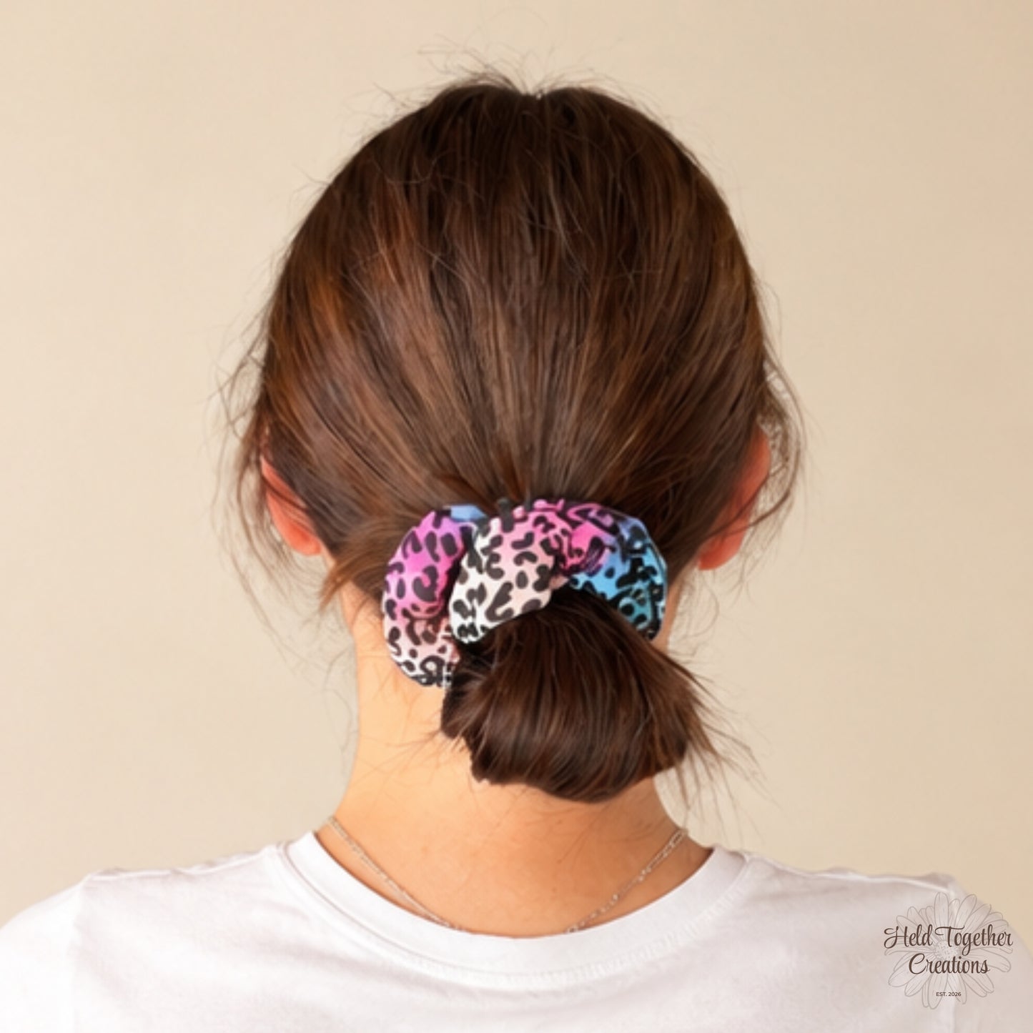 A person shown from the back wearing a leopard-print scrunchie in pink, blue, black, and white, tied around brown hair. The background is a solid light beige wall.