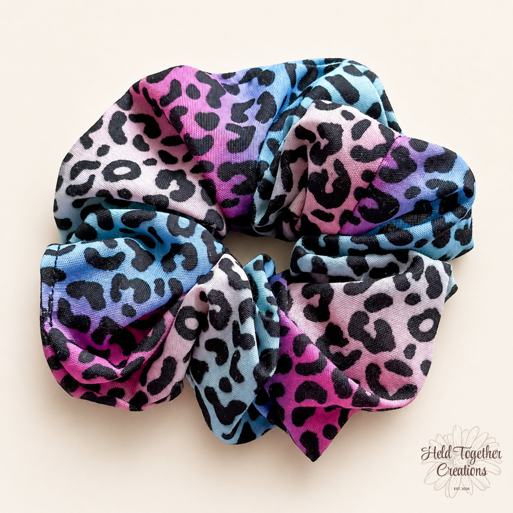 Colourful leopard print handmade scrunchie with pink, blue, purple and black tones on a soft ivory background.