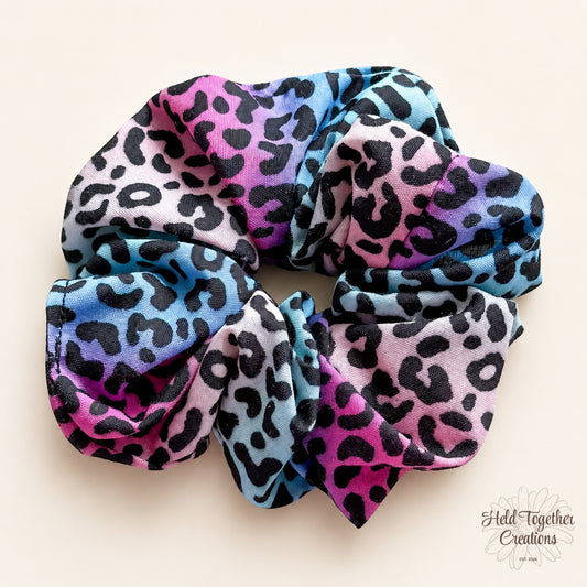 Colourful leopard print handmade scrunchie with pink, blue, purple and black tones on a soft ivory background.