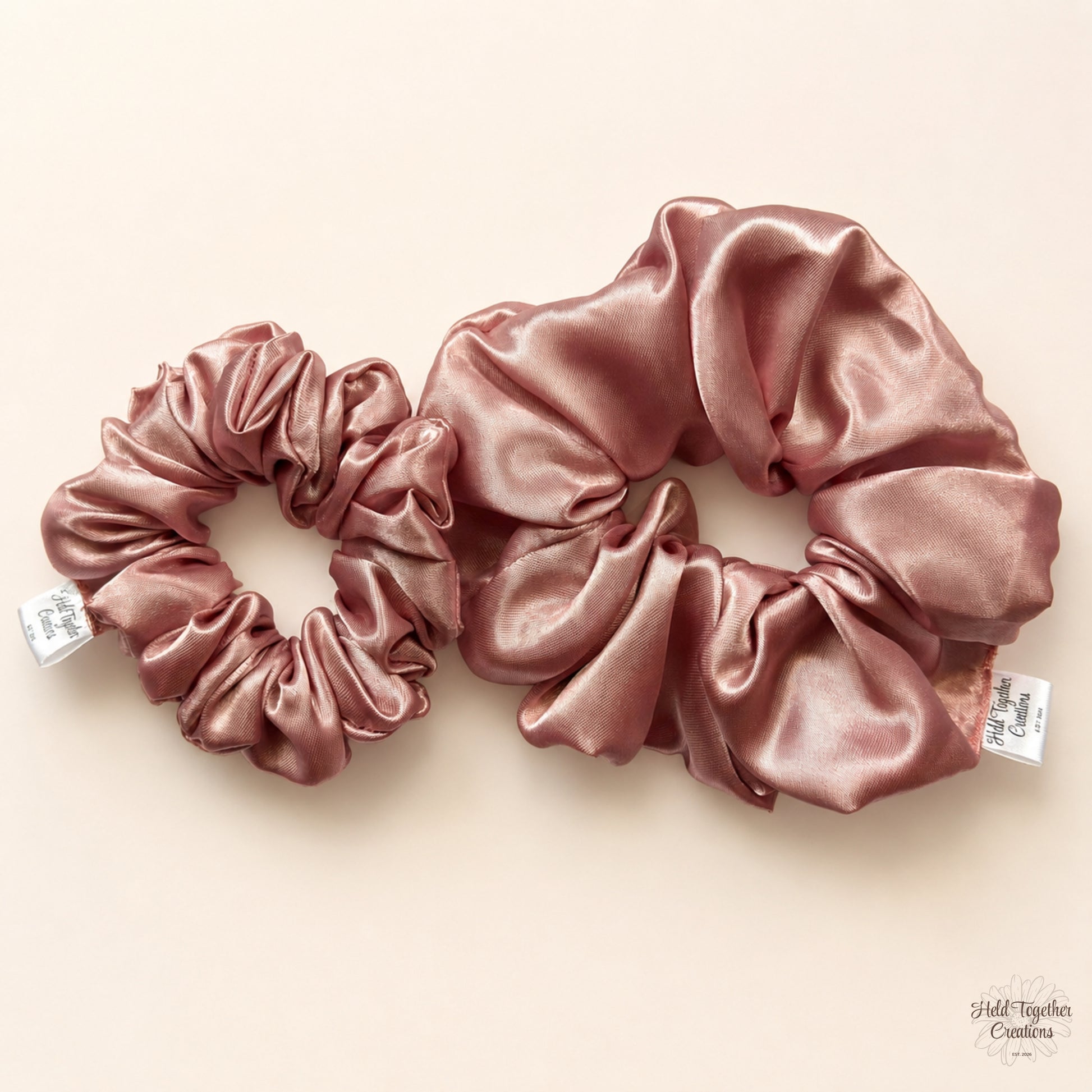 Two satin hair scrunchies in a dusty pink finish, displayed on a light beige background. One is smaller and positioned on the left, and the other is larger and positioned on the right.
