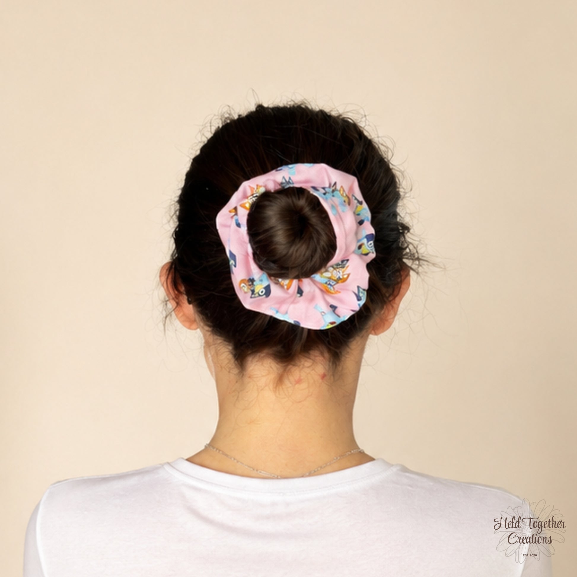 A pink fabric scrunchie with a colourful cartoon pattern, shown on a person's hair in a bun from the back against a solid beige background.