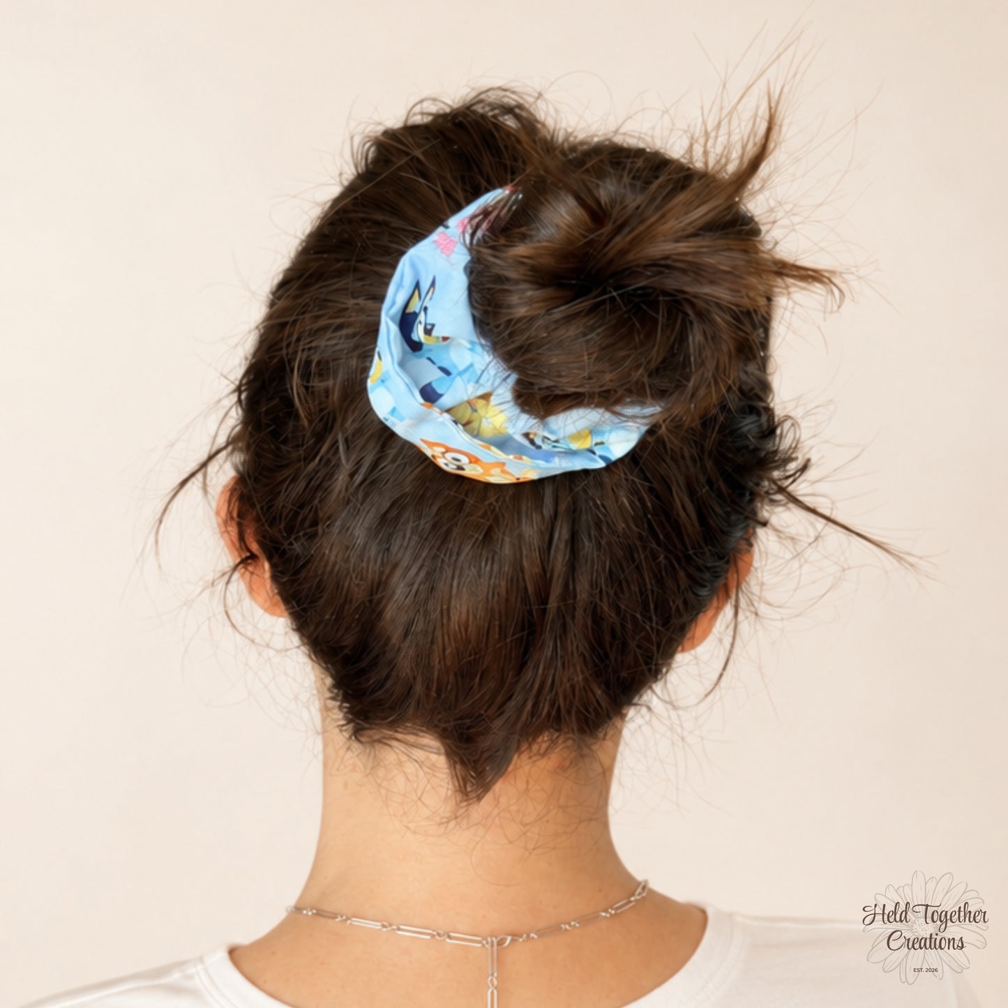 A person shown from the back wearing a light blue fabric scrunchie with a cartoon character print, holding their dark brown hair in a messy bun. The background is a solid light beige wall.