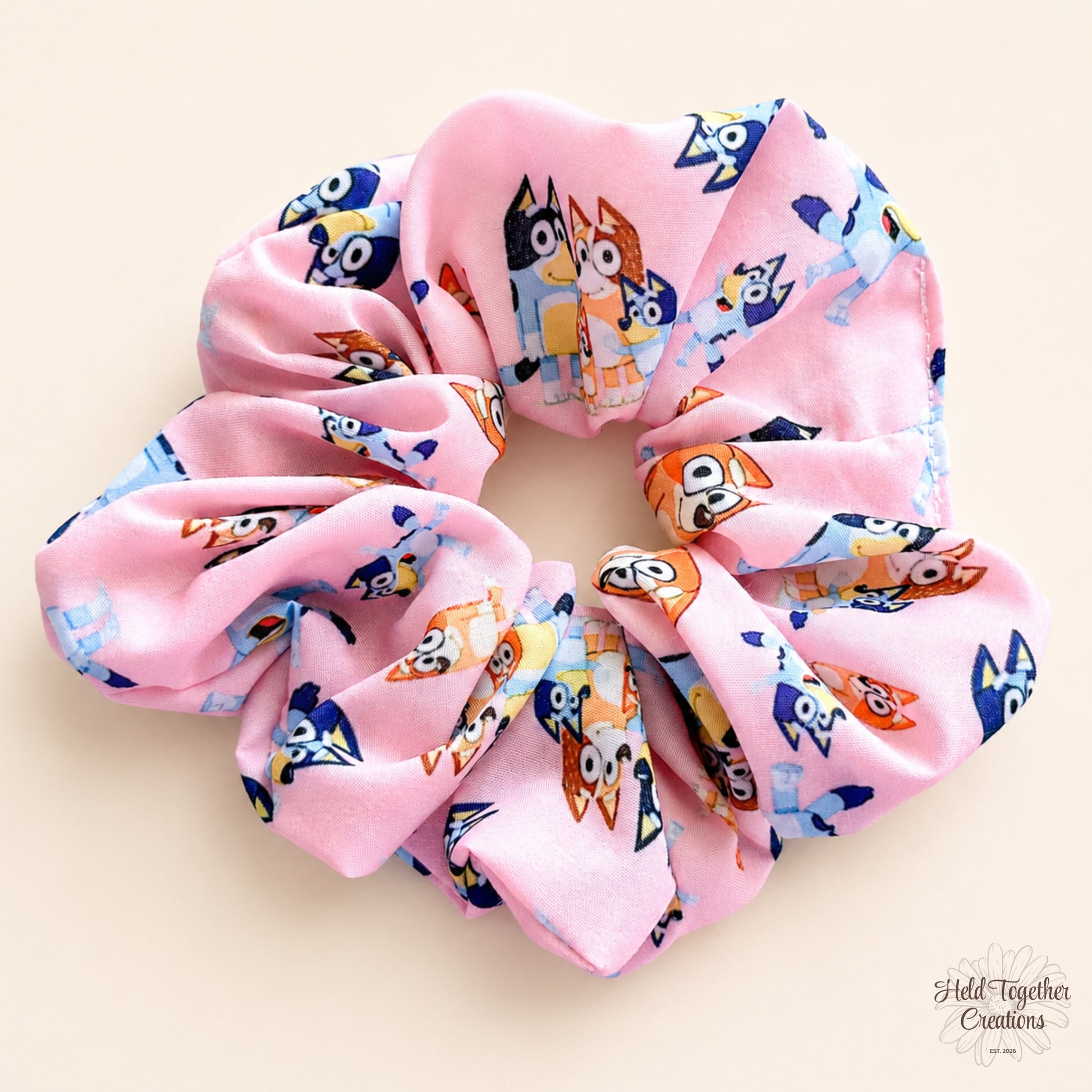 A pink fabric scrunchie featuring a colourful cartoon dog print, displayed on a light beige background.
