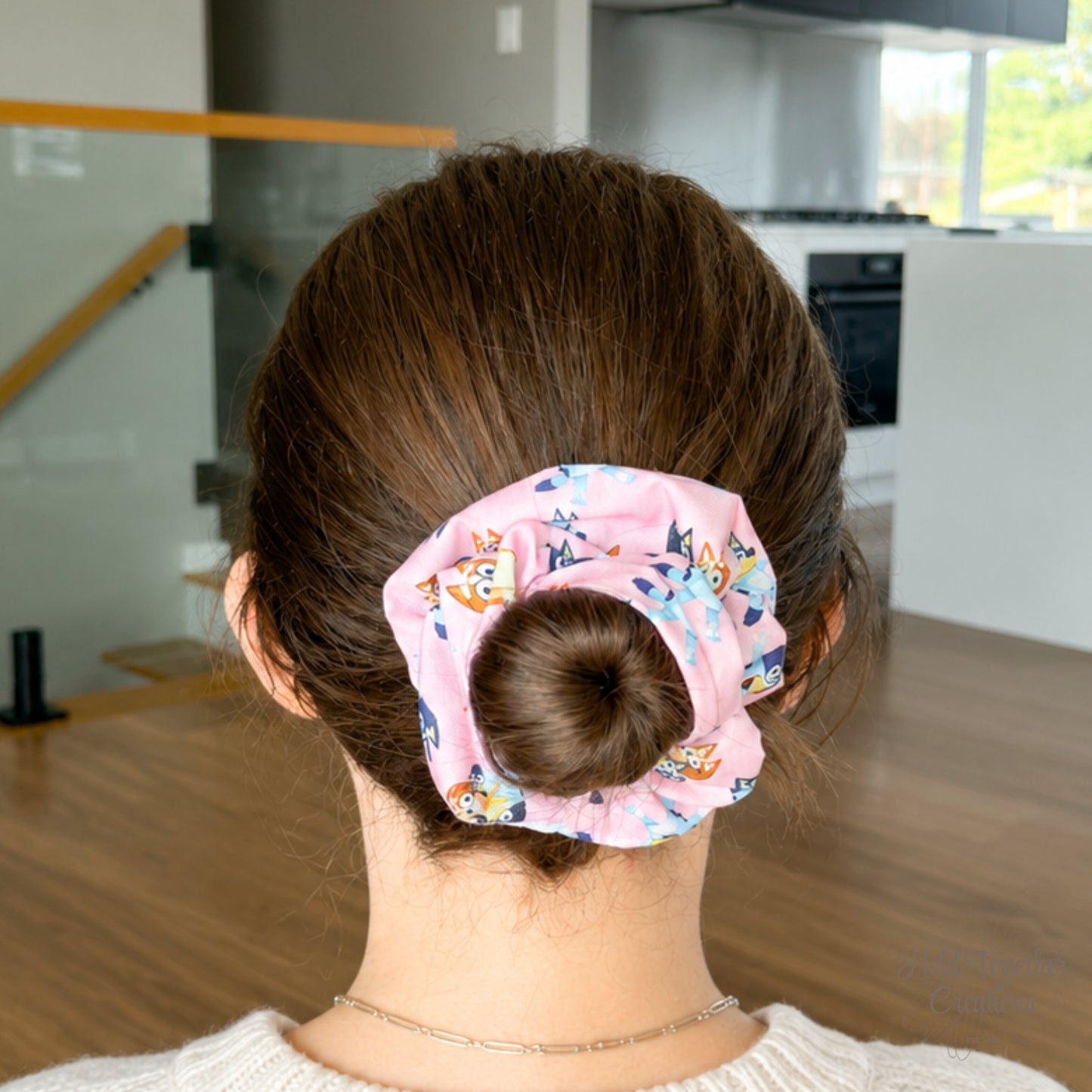 Family Pup scrunchie styled in a brown hair bun, showing the pink puppy-print fabric in a bright home setting.