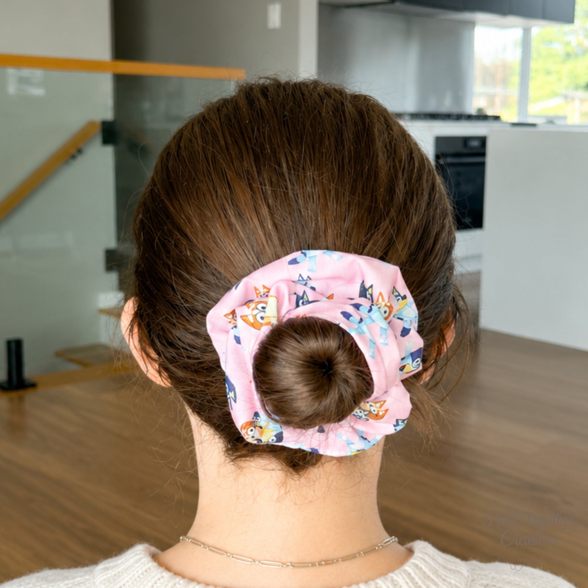 Family Pup scrunchie styled in a brown hair bun, showing the pink puppy-print fabric in a bright home setting.