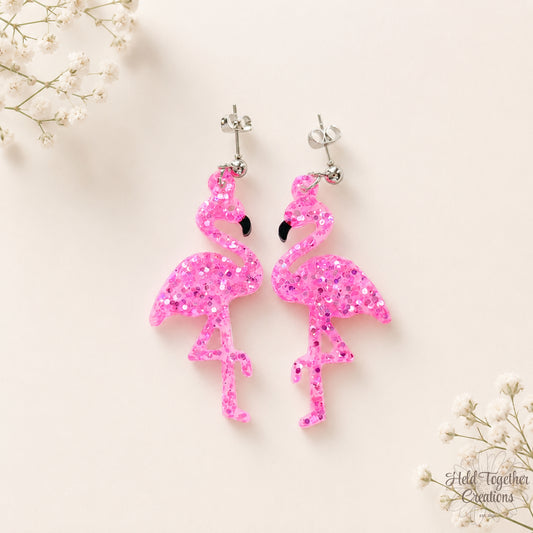 Handmade pink glitter flamingo resin earrings with silver-tone stud hardware on a soft neutral background.