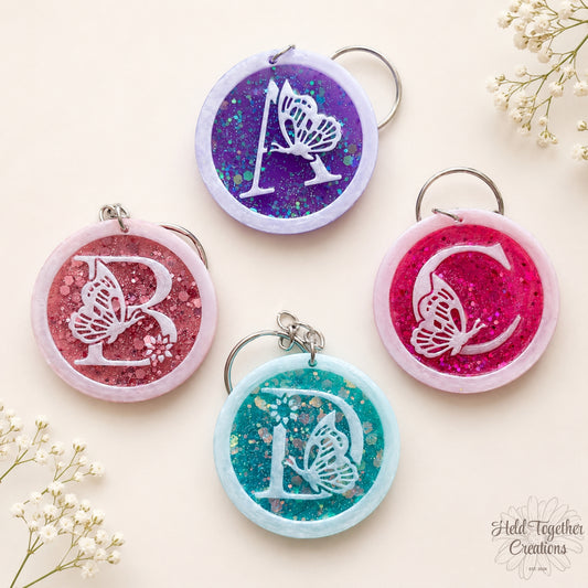 Handmade butterfly resin initial keyrings in rose gold, purple, pink and teal with glitter centres and white alphabet letters.